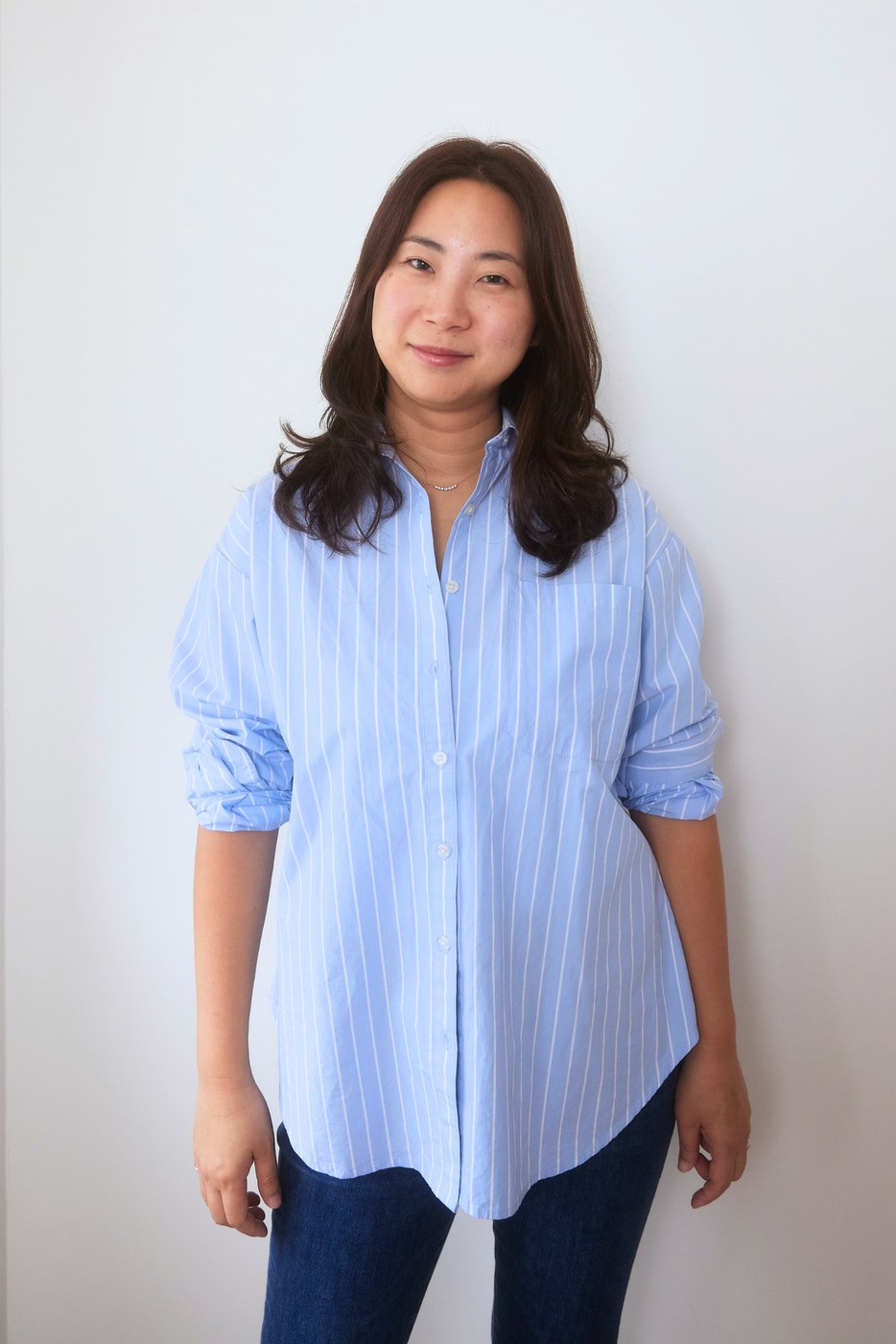 Angela Yoo standing against a white wall in a blue striped shirt